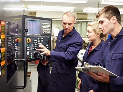 Drei Personen in Arbeitskleidung stehen vor einer Maschinensteuerung. Eine Person erklärt etwas, während die anderen zuhören und Notizen machen.