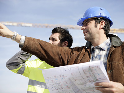 Zwei Bauarbeiter besprechen Pläne, einer zeigt auf etwas in der Ferne.