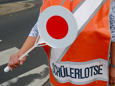 Eine Person in einer leuchtenden Warnweste hält eine Verkehrsleiteplatte mit einem roten Kreis und dem Schriftzug "SCHÜLERLOTSE".